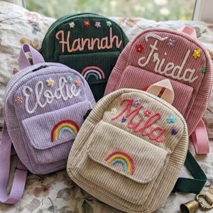 May include: Four corduroy backpacks in green, pink, lilac, and beige. Each has an embroidered name, a rainbow, and floral details. The backpacks have adjustable straps and a small front pocket.