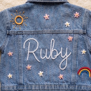 May include: A blue denim jacket featuring white embroidered text that reads Ruby in the centre. The jacket is decorated with embroidered motifs including a yellow sun, a multi-coloured rainbow, and several small pink and white flowers.