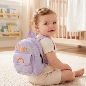 May include: A light purple corduroy mini backpack featuring the embroidered name Emmie in multi-colored script and a small rainbow patch on the front pocket. The bag has a top handle, adjustable shoulder straps, and a black zipper pull.