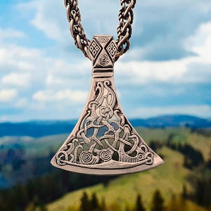 May include: A silver-colored pendant necklace featuring a detailed axe-head design with intricate knotwork. The pendant hangs from a thick, braided chain. The background shows a scenic view of rolling hills and a cloudy sky. The text "MJOLNIR WORKSHOP" is at the bottom.
