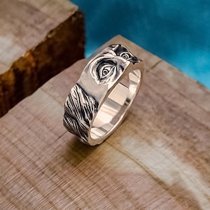 Könnte beinhalten: Ein silberner Ring mit detailliertem Gesichtsdesign, möglicherweise einer mythischen Kreatur, liegt auf einer Holzoberfläche. Das Design des Rings zeigt ein Auge und strukturierte Details. Der Ring ist vor blauem Hintergrund platziert. Der Text "MJOLNIR WORKSHOP" ist sichtbar.