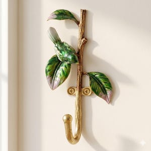 Peut inclure: Crochet mural décoratif avec un crochet doré en forme de branche, orné d'un oiseau vert et blanc et de feuilles vertes et violettes. Le crochet est conçu pour être fixé au mur.