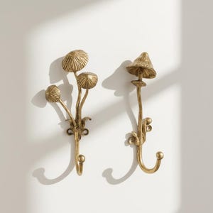 Pode incluir: Dois ganchos de parede dourados em forma de cogumelo. Um gancho apresenta um grupo de três chapéus de cogumelo, enquanto o outro tem um design de cogumelo único e mais alto. Ambos têm um gancho na parte inferior para pendurar objetos. Os ganchos são montados numa parede branca.