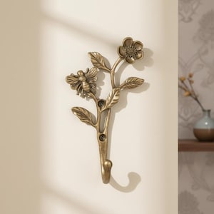 May include: A wall-mounted metal hook in a brass-toned finish, shaped like a botanical stem with leaves, a single flower at the top, and a bee perched on the side. The design includes two visible screw holes for mounting the piece to a flat surface.