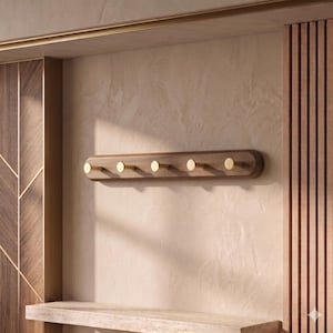 Puede incluir: Un perchero de pared horizontal con una base de madera marrón oscuro de bordes redondeados y cinco ganchos cilíndricos de latón espaciados uniformemente. El perchero presenta un acabado liso que resalta la veta natural de la madera.