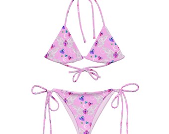 Könnte beinhalten: Pinkfarbener Bikini mit Dreieck-Oberteil und seitlich zu bindenden Höschen. Der Bikini hat einen Druck mit weißen Flügeln und rosa Herzen mit blauen Edelsteinen.