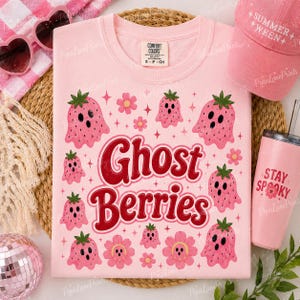 May include: A light pink t-shirt with the words "Ghost Berries" in red script, surrounded by pink strawberry ghost and flower graphics. A pink hat and tumbler with text are also visible.