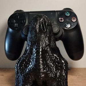 May include: A black controller stand shaped like a monster, with a textured surface. The stand is holding a black PlayStation 4 controller. The stand is on a wooden surface.