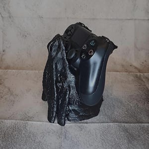 Könnte beinhalten: Ein schwarzer, texturierter Ständer in Handform, der einen schwarzen Videospiel-Controller hält. Der Controller verfügt über Standardtasten, einschließlich der Symbole Dreieck, Kreis, Kreuz und Quadrat, und steht aufrecht in den Fingern des Ständers.