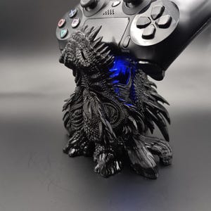 Könnte beinhalten: Ein schwarzer, texturierter Controller-Ständer in Drachenform, der einen schwarzen Gaming-Controller hält. Die Drachenfigur weist komplexe Schuppen und Stacheln auf, wobei ein helles blaues Licht aus dem Brustbereich leuchtet und eine stabile Basis bietet.