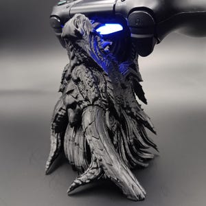 Könnte beinhalten: Ein schwarzer, 3D-gedruckter Controller-Ständer in Form eines Drachenkopfes mit detaillierten Schuppenstrukturen und spitzen Hörnern. Der Ständer hält einen schwarzen kabellosen Gamecontroller, wobei ein blaues Licht unter dem Gerät leuchtet.