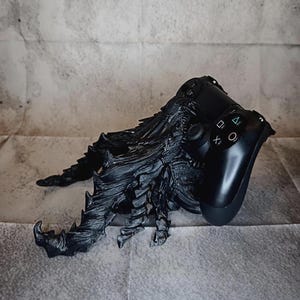 Könnte beinhalten: Ein schwarzer, texturierter Controller-Ständer in Form einer Kreatur mit mehreren stacheligen, klauenartigen Gliedmaßen. Der Ständer ist so konzipiert, dass er einen schwarzen Gaming-Controller hält, der sicher in der dunklen, gezackten Form ruht.