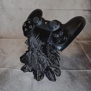 Könnte beinhalten: Ein schwarzer Gaming-Controller ruht auf einem schwarzen Ständer in Drachenform. Die Drachenfigur weist komplexe, texturierte Schuppen und spitze Details am ganzen Körper auf und bietet eine stabile Basis, um den Controller sicher zu halten.