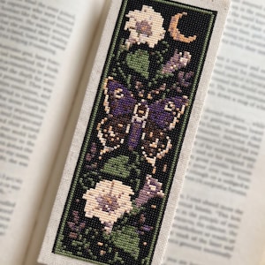 Puede incluir: Un marcapáginas rectangular de punto de cruz con una polilla morada central rodeada de flores blancas, hojas verdes y una pequeña luna creciente. El diseño se presenta sobre un fondo negro con un borde verde en una base de tela clara.