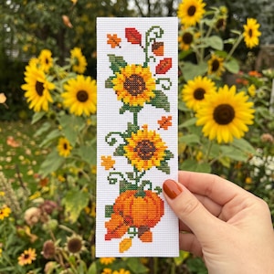 May include: A rectangular white bookmark featuring a cross-stitch pattern of two yellow sunflowers, green leaves, a bright orange pumpkin, and small scattered autumn leaves in red, orange, and yellow colors arranged in a vertical design.