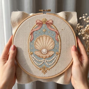 May include: A cross-stitch embroidery in a round wooden hoop featuring a central open seashell with a white pearl inside. The design is framed by a gold border, a pink ribbon at the top, and draped white pearl strings on a light blue background.