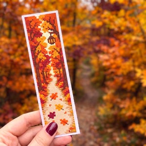May include: A rectangular cross-stitch bookmark featuring a vertical path winding through a dense forest of autumn trees. The design displays vibrant shades of orange, red, yellow, and brown, with scattered fallen leaves on the path and a lantern.