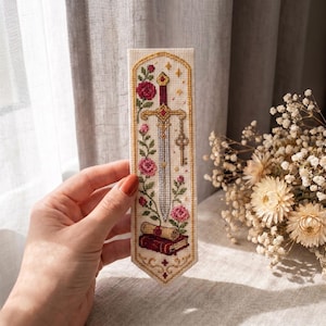 May include: A rectangular fabric bookmark featuring a cross-stitch design of a central sword, a hanging key, pink roses, and a scroll resting on a red book. The embroidery is framed by a gold border on a cream-colored background with a pointed base.