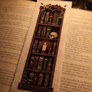 Peut inclure: Un marque-page rectangulaire en point de croix sur tissu beige clair représentant une bibliothèque en bois brun foncé. Les étagères sont remplies de divers livres, d'un petit crâne humain blanc et d'une bougie allumée au centre.