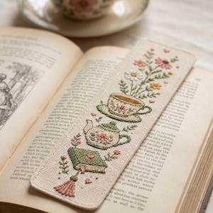 Könnte beinhalten: Ein rechteckiges Lesezeichen aus Stoff mit Kreuzstichstickerei, die ein grünes Buch mit rosa Quaste, eine Teekanne und eine blumige Teetasse zeigt. Das Lesezeichen liegt auf einem offenen Buch, im Hintergrund steht eine passende Teetasse.