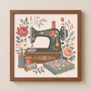 May include: A cross-stitch embroidery in a wooden frame depicting a dark green vintage sewing machine decorated with pink roses. The scene includes three spools of thread in blue and pink, a folded piece of floral fabric, and surrounding greenery.