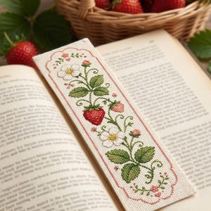 Könnte beinhalten: Ein rechteckiges Lesezeichen aus Stoff mit einem Kreuzstichmuster aus Erdbeerpflanzen mit weißen Blüten, grünen Blättern und roten Beeren. Das Design ist von einem gewellten, rosa gestickten Rand auf cremefarbenem Webstoff umgeben.