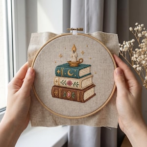 May include: A circular wooden embroidery hoop containing a piece of light beige fabric with a cross-stitch design of a stack of three books topped with a lit candle. The books feature decorative motifs including a crescent moon, a rose, and stars.