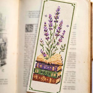 May include: A rectangular cross-stitch bookmark on a cream-colored fabric base. The design features a stack of three books in purple, green, and brown, with a cluster of purple lavender sprigs and green stems emerging from the top book.