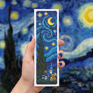 Puede incluir: Un marcapáginas rectangular con un patrón de punto de cruz inspirado en La noche estrellada, que muestra una luna creciente amarilla, estrellas brillantes, un ciprés verde oscuro y un pequeño pueblo con edificios azules y amarillos.
