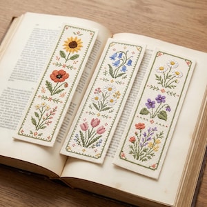Peut inclure: Trois marque-pages rectangulaires présentant des motifs floraux détaillés au point de croix sur un tissu crème. Les designs incluent des tournesols, coquelicots, campanules, marguerites, tulipes et violettes, bordés de cadres verts et de motifs géométriques.