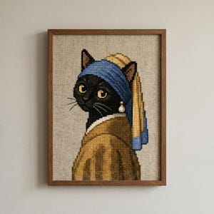 May include: A cross-stitch embroidery on a light beige fabric featuring a black cat wearing a blue headscarf and a tan garment. The cat has a single white pearl earring on its right ear. The entire piece is framed in a simple brown wooden frame.