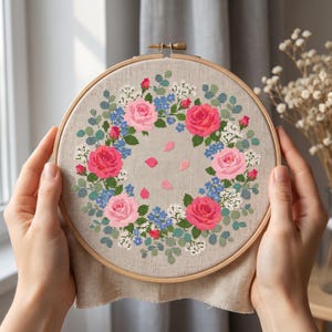 May include: A circular wooden embroidery hoop containing a cross-stitch pattern of a floral wreath. The design features pink and red roses, small blue flowers, white blossoms, and green leaves on a light beige fabric background.