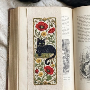 May include: A rectangular fabric bookmark featuring an embroidered design of a black cat resting on a green book, surrounded by red poppy flowers, yellow blossoms, and green vines, all enclosed within a decorative gold-toned rectangular border.