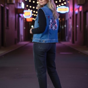 Puede incluir: Un chaleco vaquero azul claro con un diseño de calavera y rosas en la espalda. Se lleva sobre una camiseta negra de manga larga y vaqueros grises oscuros. La persona lleva botas negras. El fondo es una calle de la ciudad de noche.