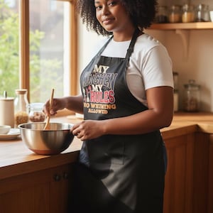 Puede incluir: Un delantal negro con el texto "MY KITCHEN MY RULES NO WHINING ALLOWED" es usado por una persona en una cocina. La persona está revolviendo un cuenco con una cuchara de madera. La cocina tiene armarios de madera y una ventana.