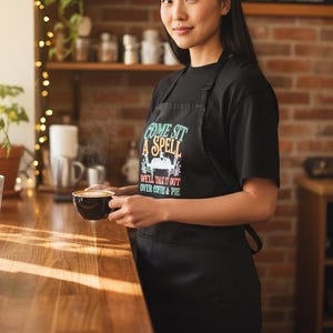 Puede incluir: Una persona con un delantal negro con el texto "COME SIT A SPELL WELL TALK IT OUT OVER COFFEE & PIE" sosteniendo una taza de café. El fondo incluye una encimera de madera, una pared de ladrillo y estantes.
