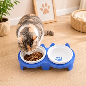 Pode incluir: Um suporte duplo azul para animais de estimação com duas taças de cerâmica branca removíveis. Uma taça está cheia de ração seca castanha, enquanto a outra apresenta um gráfico de pata azul no fundo. O suporte tem formas de orelhas de gato.