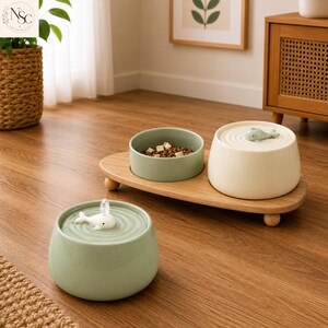 May include: A pet feeding station featuring a light green ceramic food bowl and a cream-coloured ceramic water fountain on a wooden stand, plus a separate light green ceramic water fountain, both topped with small white whale figurines.