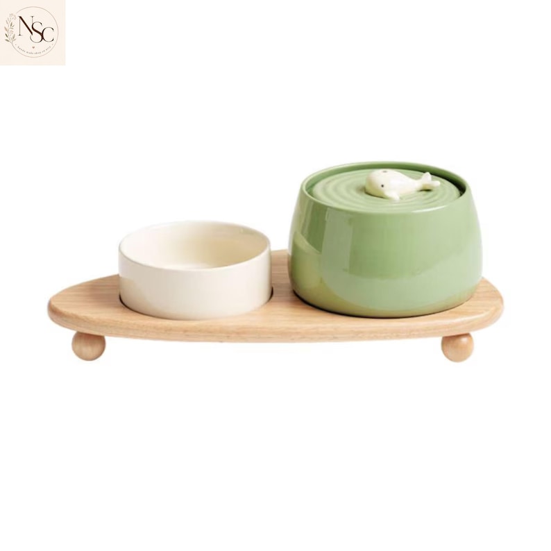 May include: A pet feeding station featuring a light wood base with three small ball feet. It holds a plain cream-coloured ceramic bowl and a larger sage green ceramic bowl topped with a lid featuring a small white whale figurine on a ripple design.