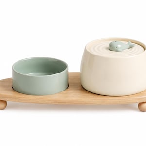 May include: A light green ceramic bowl and a cream-coloured ceramic container with a lid featuring a small whale figurine, both resting on a light-toned wooden tray with three rounded feet. The lid has a circular ripple pattern around the whale.
