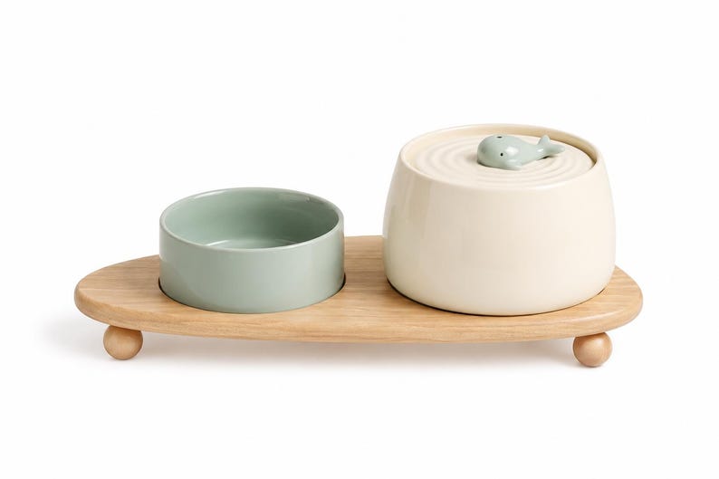 May include: A light green ceramic bowl and a cream-coloured ceramic container with a lid featuring a small whale figurine, both resting on a light-toned wooden tray with three rounded feet. The lid has a circular ripple pattern around the whale.