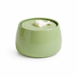May include: A sage green ceramic pet water fountain with a smooth, rounded exterior. The top surface features a concentric ripple pattern and a small, white, sculpted whale figurine positioned in the centre, serving as the water spout outlet.