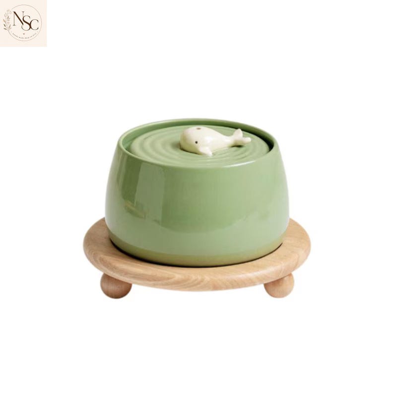 May include: A sage green ceramic bowl with a smooth, rounded silhouette. The top features a concentric ripple pattern and a small, white, sculpted whale figurine. The bowl rests on a circular, light-toned wooden base with three small feet.
