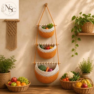 May include: Three-tiered, hanging crochet baskets in orange and white, filled with fresh produce. The baskets are suspended from a wooden dowel and rope. Baskets of fruit and vegetables are on a wooden surface. A macrame wall hanging is on the left.