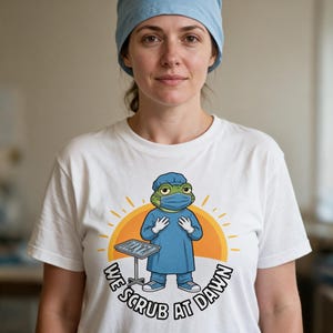 Op de afbeelding: Een wit t-shirt met een afbeelding van een groene kikker in blauwe operatiekleding, met een mutsje en een mondkapje. De kikker staat naast een metalen dienblad met chirurgische instrumenten. Onder de afbeelding staat de tekst "WE SCRUB AT DAWN".