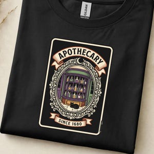 May include: A black t-shirt featuring a graphic print of an apothecary shop interior with shelves of colourful bottles and a black cat. The design is enclosed in an ornate frame with banners reading APOTHECARY and SINCE 1680 in black text.