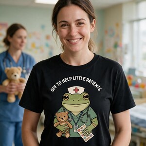 May include: A black t-shirt featuring a graphic of a green frog dressed as a medical professional wearing a nurse cap with a red cross, a stethoscope, and holding a brown teddy bear and a sheet of colorful stickers with the text OFF TO HELP LITTLE PATIENTS.