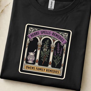 May include: A black t-shirt featuring a graphic print of three arched columns. The design includes a mortar and pestle, a sprig of rosemary, and lavender. Text reads: HERBS SPELLS REMEDIES and OWENS FAMILY REMEDIES. The shirt has a Gildan brand label.