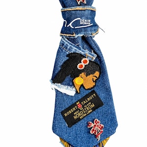 May include: A blue denim necktie featuring a red crystal butterfly brooch, a white embroidered logo reading DENIM WOMAN, a black patch with the text ROBERT TALBOTT NORDSTROM, an embroidered profile of a person, and a red crystal flower brooch.