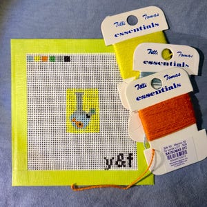 May include: A square needlepoint canvas with a lime green border featuring a small central graphic of a blue and orange water pipe on a yellow background, with the text y&f in the corner, accompanied by three Tilli Tomas essentials thread spools.